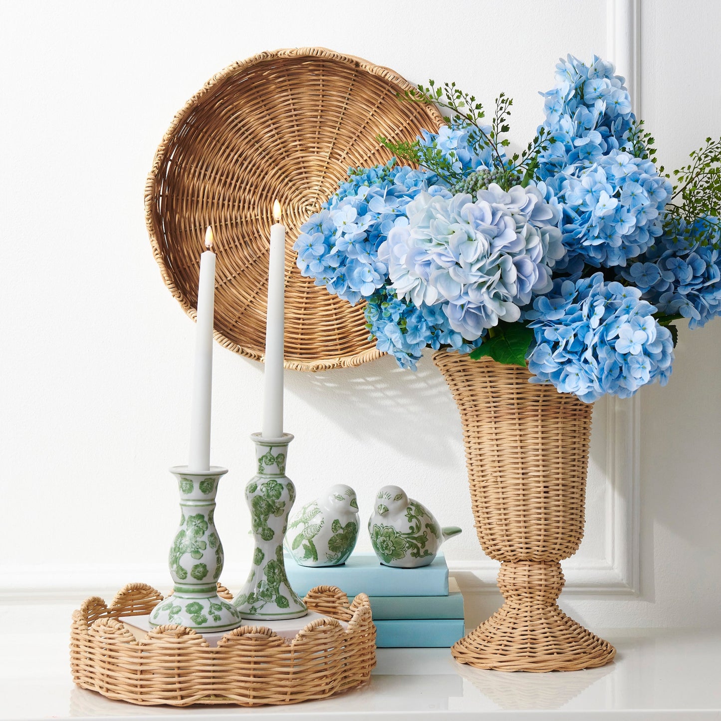 Decorative setting with blue flowers in a wicker vase, candles, and a woven basket on a white surface.