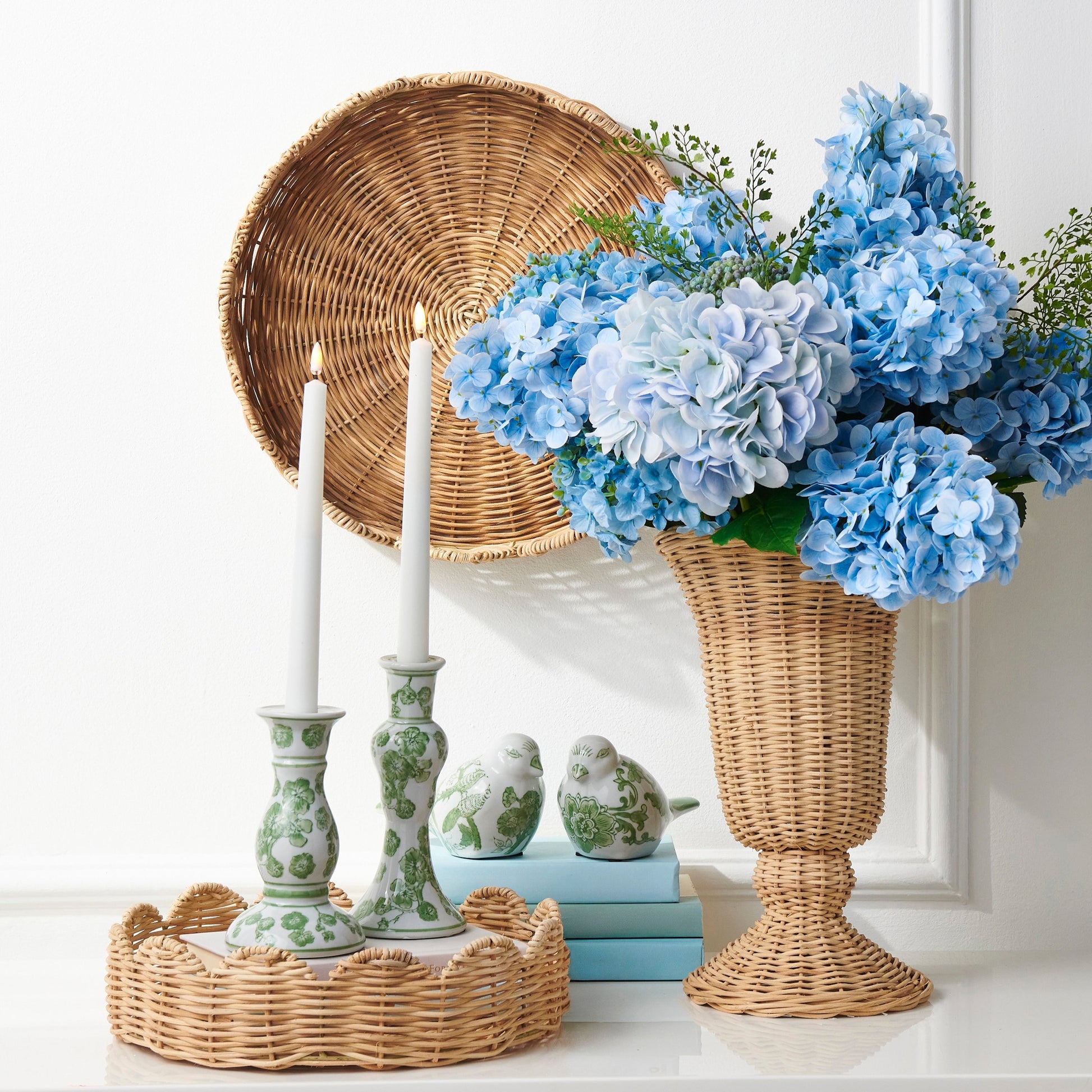 Decorative setting with blue flowers in a wicker vase, candles, and a woven basket on a white surface.