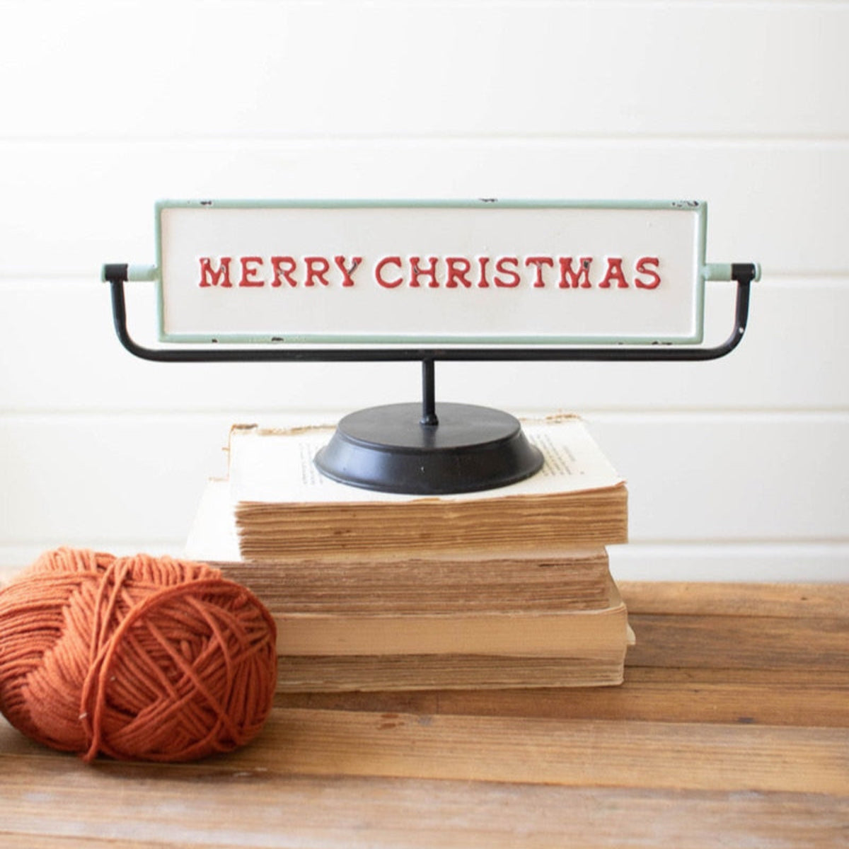 A festive, dual-sided enamel flip sign displaying 'Merry Christmas' on one side, placed on a stack of books with a ball of yarn beside it, against a wooden backdrop.