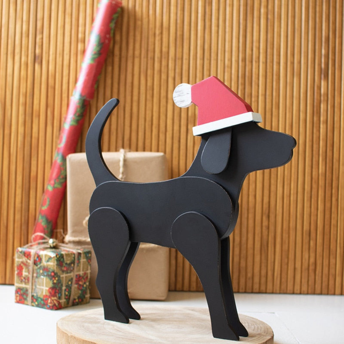 A wooden tabletop Christmas decoration featuring a black Labrador retriever wearing a Santa hat, standing next to a wrapped gift and a Christmas tree.