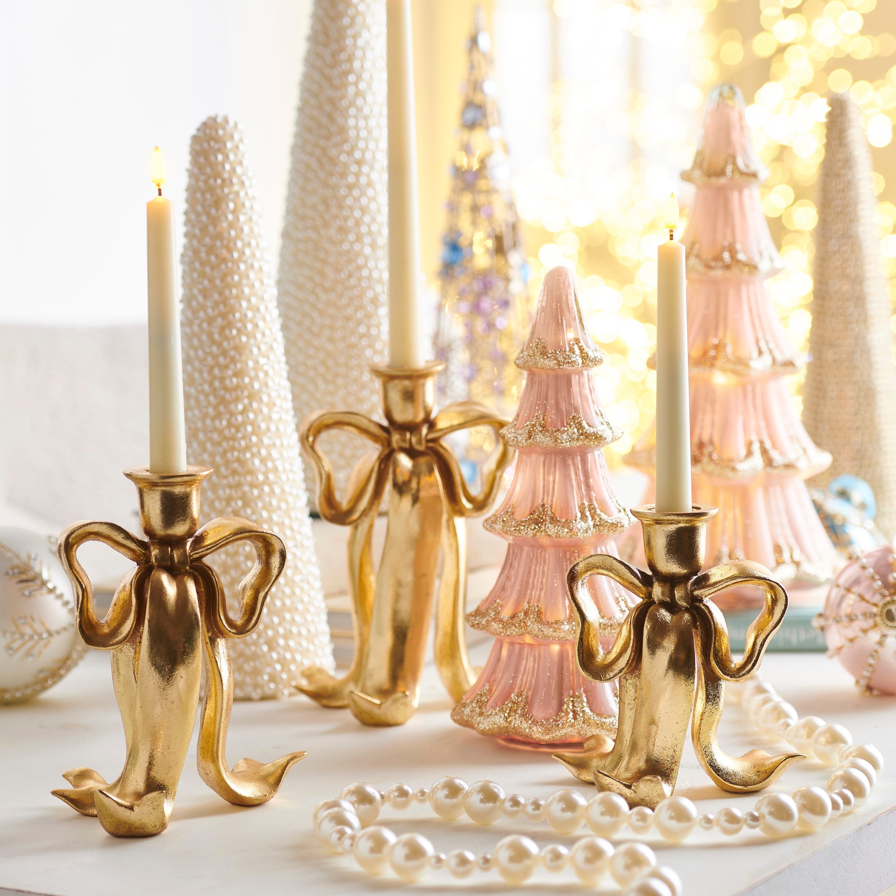 A set of three golden bow-shaped candlestick holders displayed with decorative items in the background, including Christmas trees and lights.