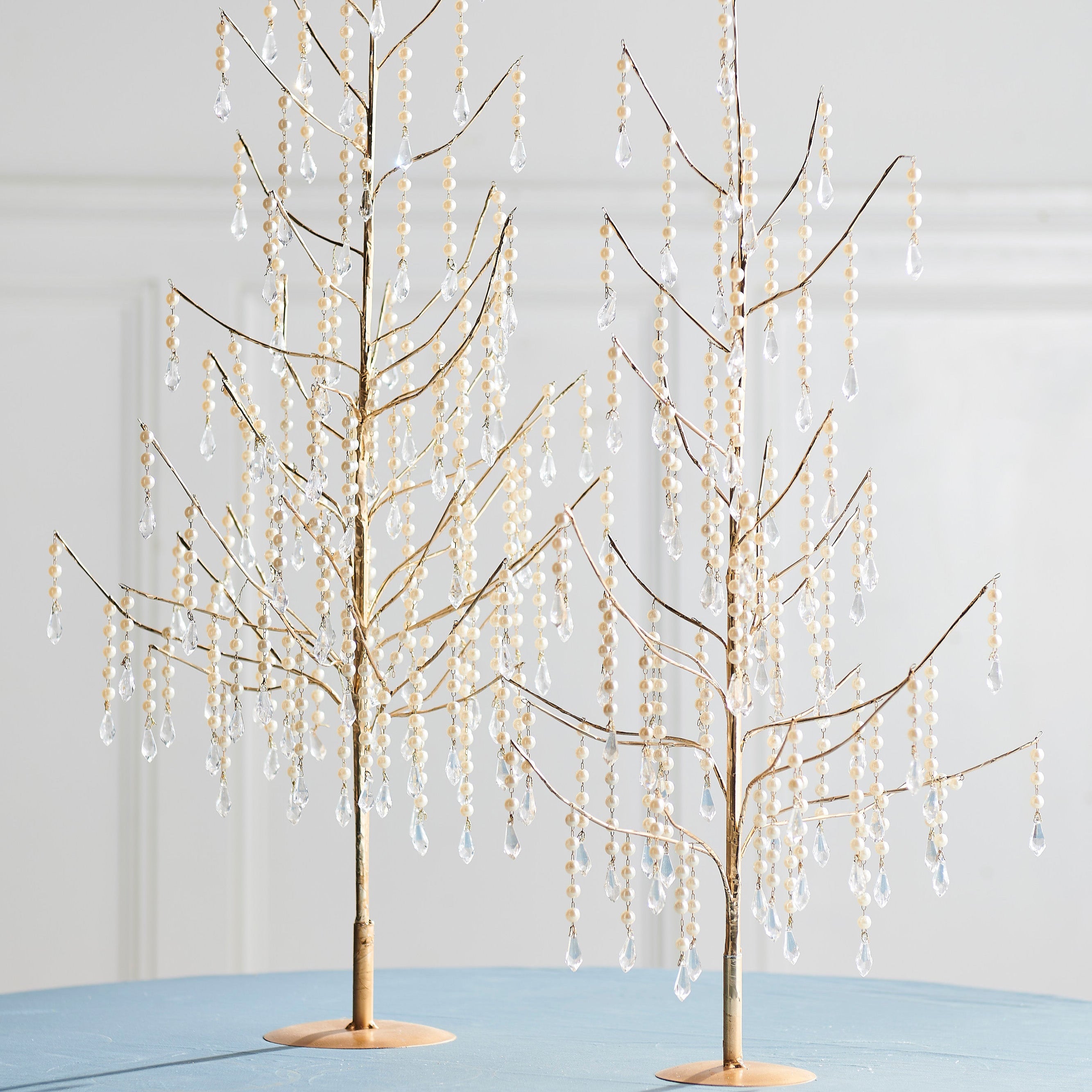 Two decorative trees with a gold finish and sparkling pearl and jewel accents, displayed on a table with a light blue cover.
