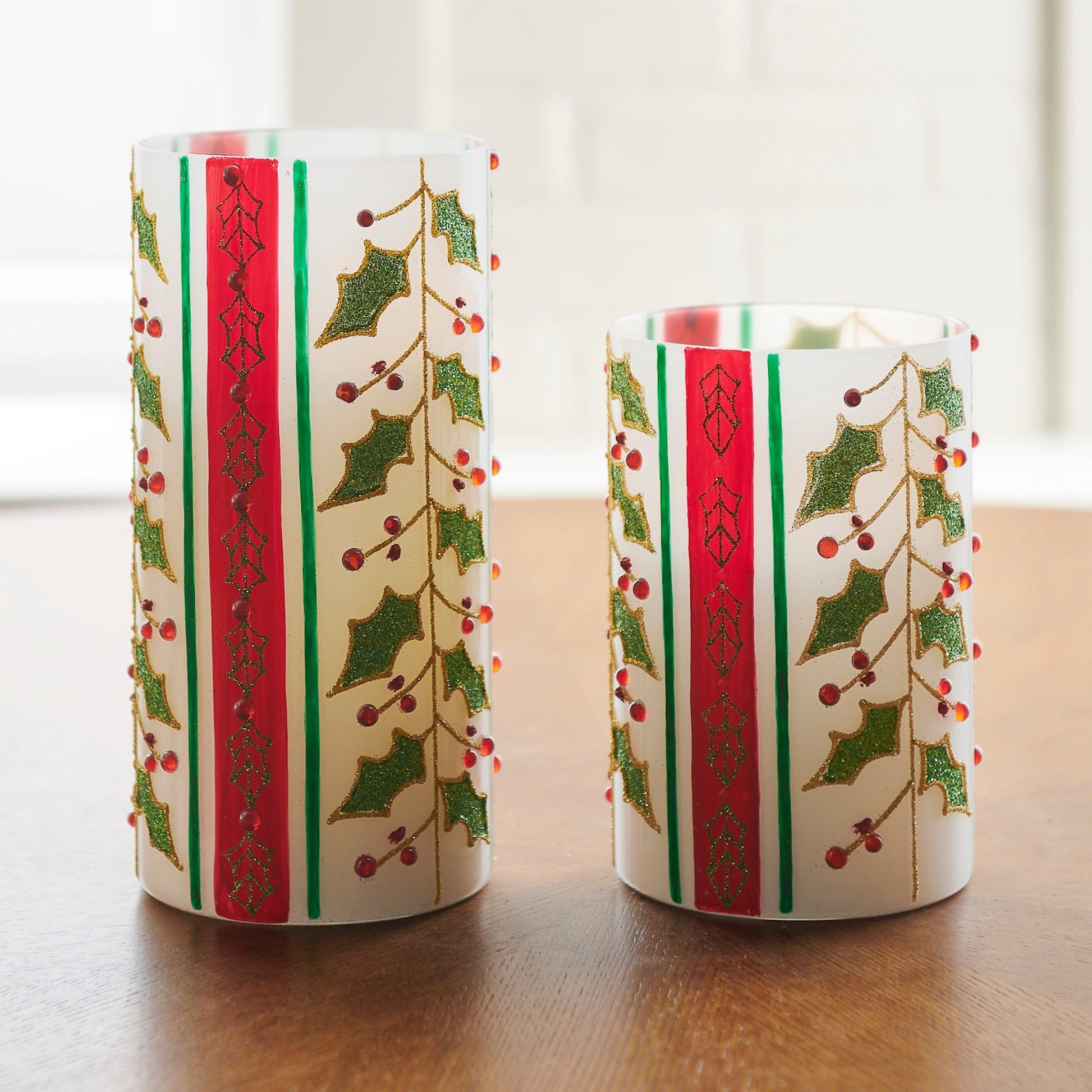 Two decorative hurricanes with holly design on a wooden surface