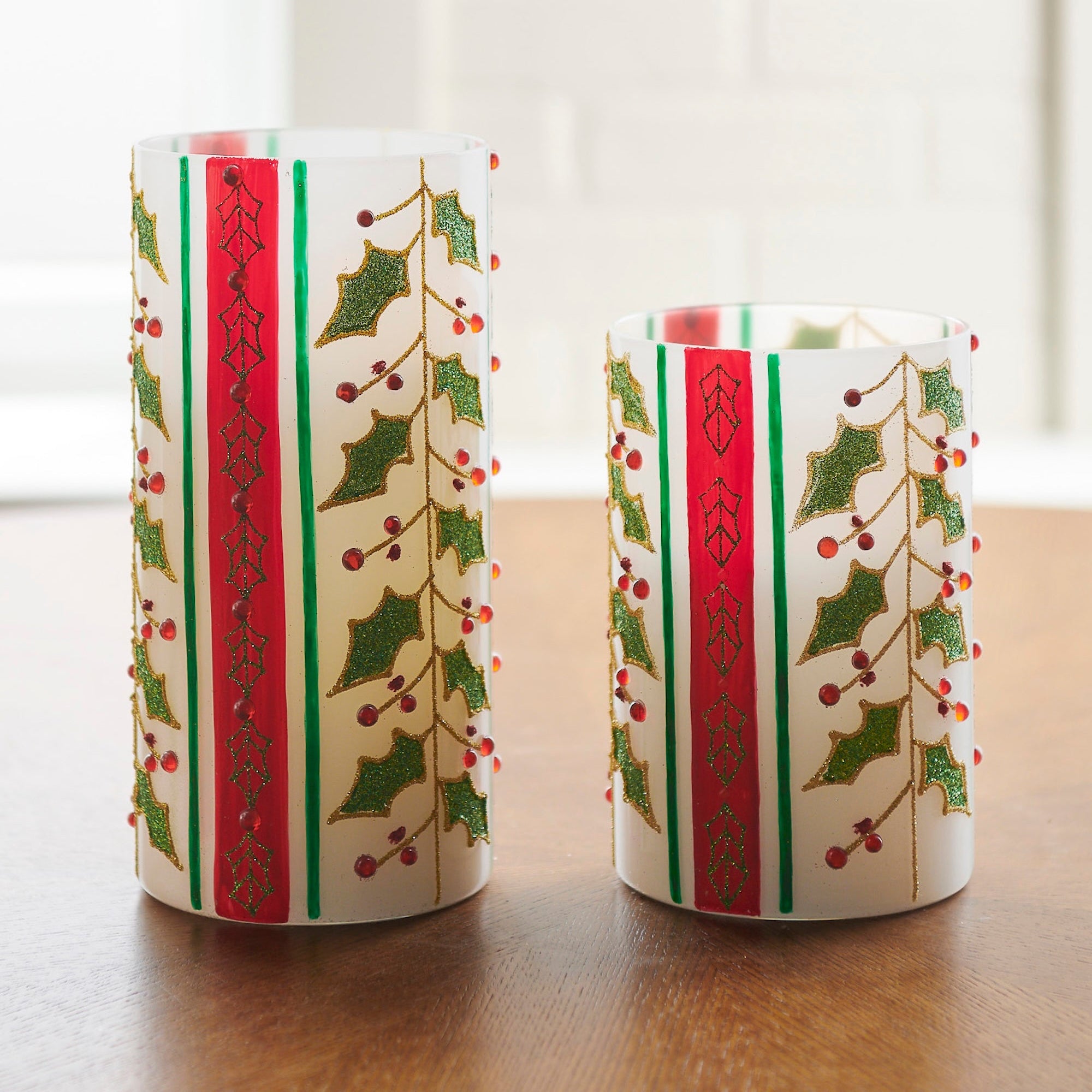 Two decorative hurricanes with holly design on a wooden surface