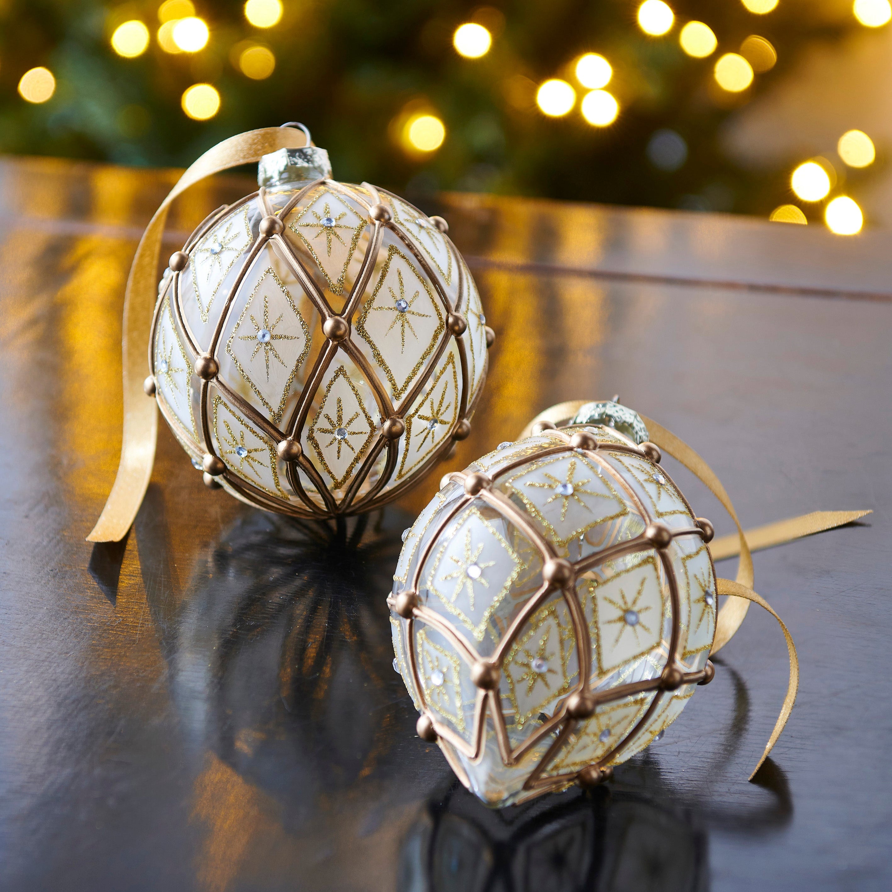 Two glass Christmas ornaments with a gold deco design and North Star motif, displayed on a reflective surface with blurred Christmas lights in the background.