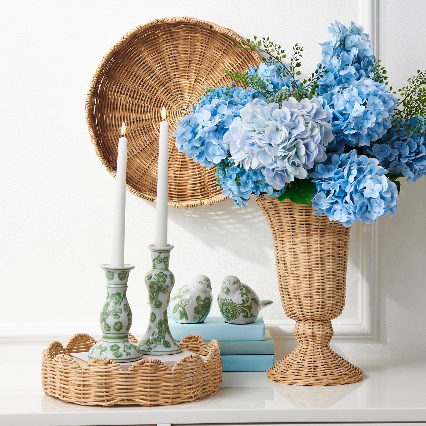 Decorative setting with blue hydrangeas in a wicker vase, candles, and a woven basket on a white surface.