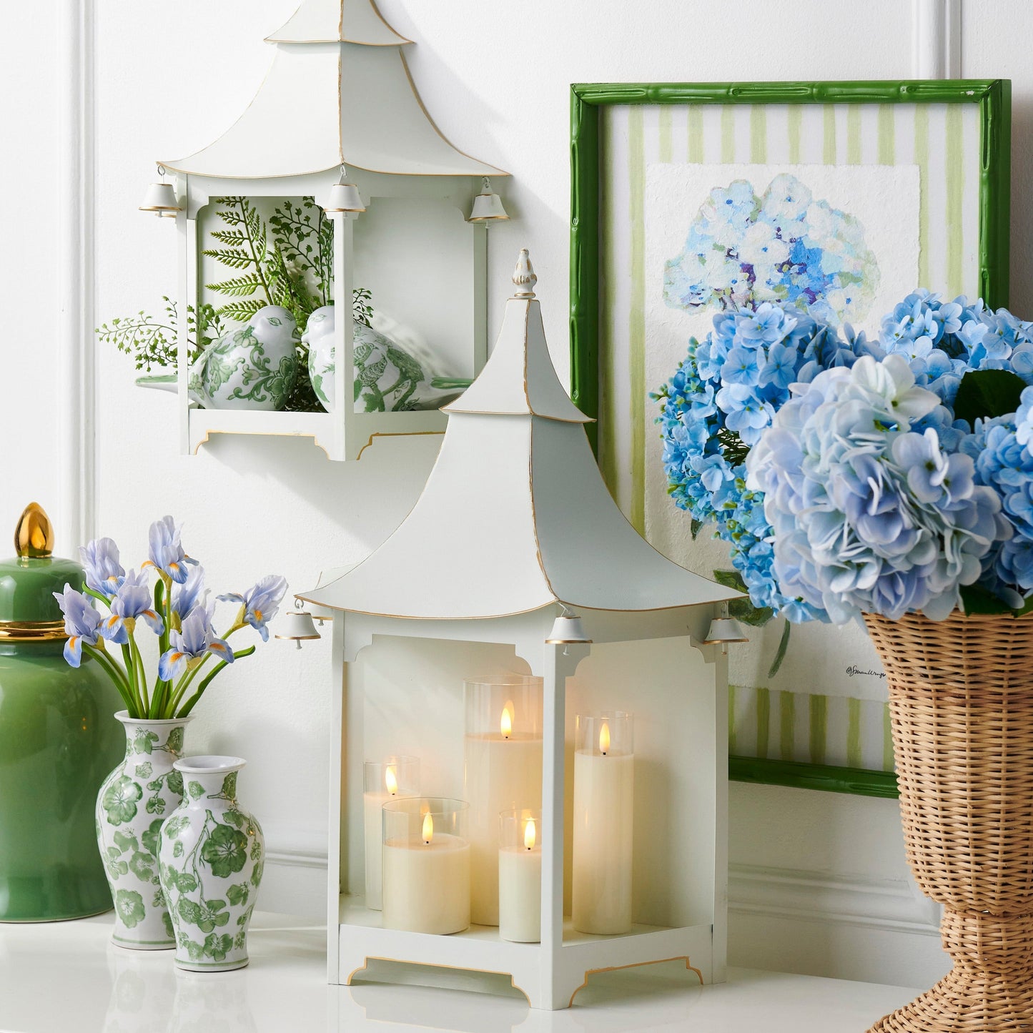Decorative setting with white pagoda lantern, candles, vases, and a basket of flowers on a white surface.