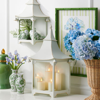 Decorative setting with white pagoda lantern, candles, vases, and a basket of flowers on a white surface.